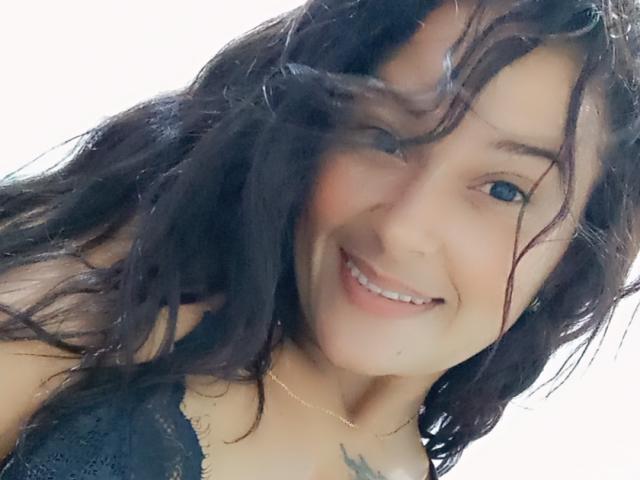 TefhaStar con capelli neri ondulati, che sorride, con occhi azzurri, indossa una camicetta in pizzo, mostra un piccolo tatuaggio sul petto, sfondo bianco, luce soffusa, capelli leggermente spettinati..