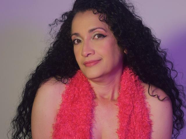 TitaNaughty with curly black hair, pink fluffy scarf, and subtle smile, showing her tits in a pink fluffy scarf. She's a cute, chubby, fair-skinned woman.