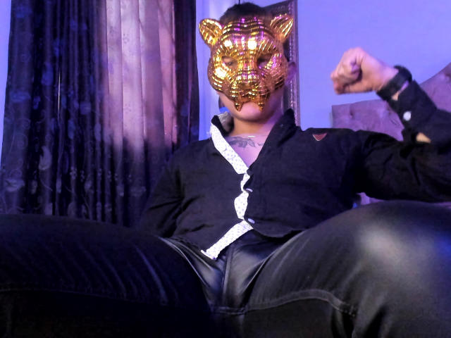 TimThomis, con una máscara de tigre dorada, una camisa negra y pantalones de cuero negro, flexiona su brazo derecho, con un tatuaje visible en el cuello, y lleva una pulsera en el brazo izquierdo..