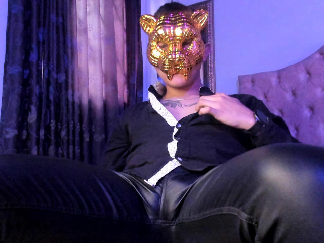 TimThomis, con una máscara de tigre dorada, una camisa negra con detalles blancos y pantalones de cuero negro, sentado con las piernas separadas, con una mano en la camisa, en una habitación con poca luz..