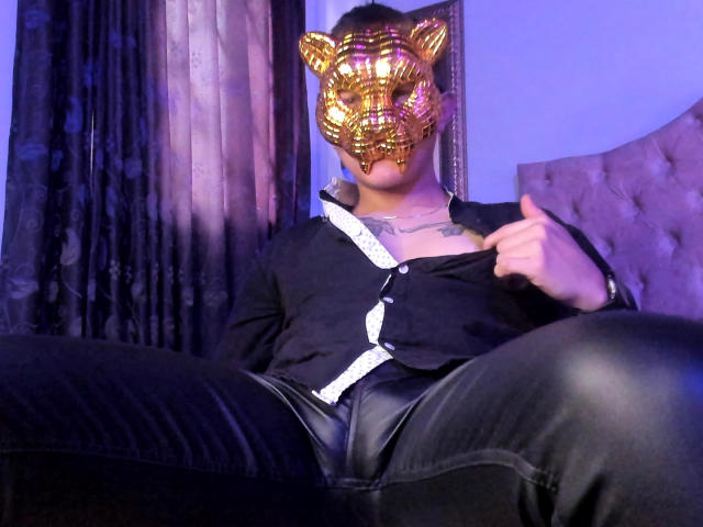 TimThomis trägt eine goldene Tiger-Maske, ein schwarzes Hemd und Lederhosen, sitzt mit gespreizten Beinen, die rechte Hand auf dem Hemd, blau-violettes Licht, kurze, dunkle Haare, Tattoo auf der Brust..