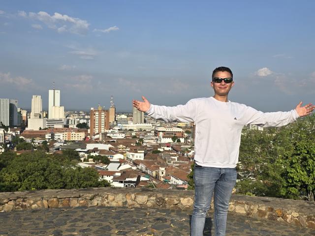 TimThomis está de pie con los brazos extendidos, vistiendo una camisa blanca de manga larga, jeans azules y gafas de sol negras, en un mirador de adoquines con una ciudad y árboles verdes al fondo..