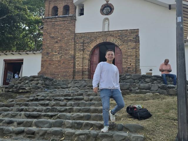 TimThomis, with tits covered by a white long-sleeve shirt, jeans, and white sneakers, stands on stone steps in front of a brick building with a red door.