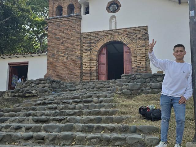 TimThomis with a white long-sleeve shirt, blue jeans, and sneakers, standing on stone steps, making a peace sign, with a backpack on the ground.