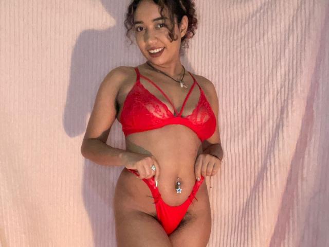 OctaviaSteel with curly brown hair, smiling, wearing red lace bra and matching thong, showing her tits and pussy, standing against pink fabric, belly button ring visible.