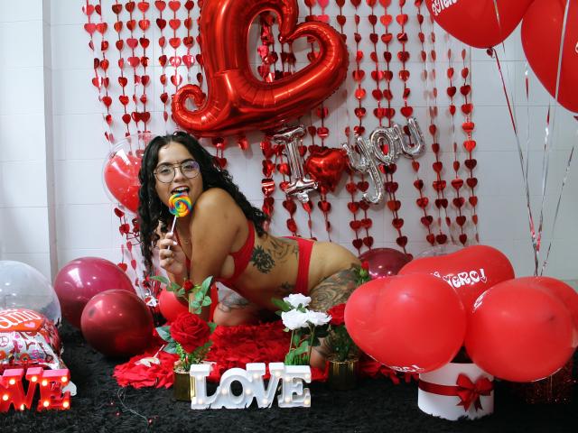 TinaFiery, met krullend zwart haar, een bril en rode lingerie, houdt een lolly vast, knielt op een zwart tapijt, omringd door rode ballonnen, harten en decoraties met de tekst "Ik hou van jou". Haar billen en tatoeages zijn zichtbaar..