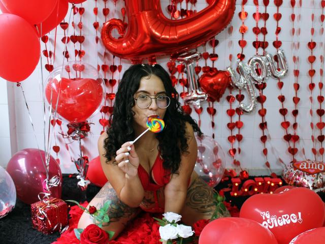 TinaFiery, met lang zwart haar, een bril, een rode beha en tatoeages, zuigt aan een kleurrijke lolly, omringd door rode ballonnen, hartdecoraties en ballonnen met de tekst "Ik hou van jou"..