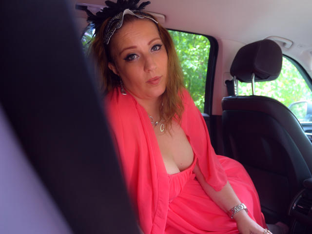 VanessaSquirtt with blonde hair, wearing a black lace headpiece, pink dress, and necklace, sitting in a car seat, looking at the camera with pouty lips. Boobs visible.