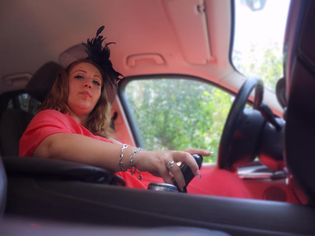 VanessaSquirtt in a red dress, black hat with feathers, and silver bracelet, gripping the car steering wheel, looking forward, with her hand on the gearshift.