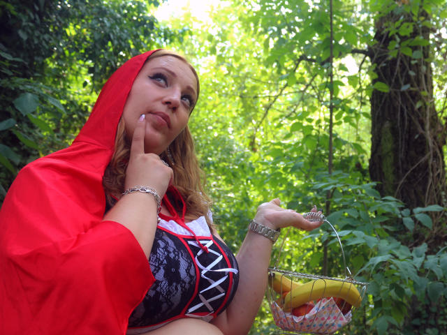 VanessaSquirtt with blonde hair, red hood, large tits in black lace-up corset, holding fruit basket, finger on chin, green forest background, looking up, silver bracelet, wristwatch.