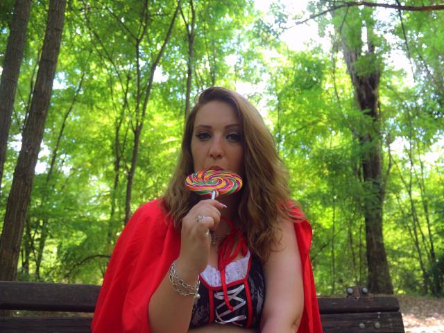 VanessaSquirtt with wavy brown hair, red cape, and black corset, sucking on a colorful swirl lollipop, sitting on a wooden bench in a sunny forest.