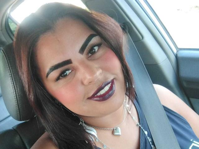 TaisaPoderosa's face in a car, wearing dark purple lipstick, heart necklace, and seatbelt, with brown hair, thick eyebrows, and a slight smile. Her tits are partially visible.