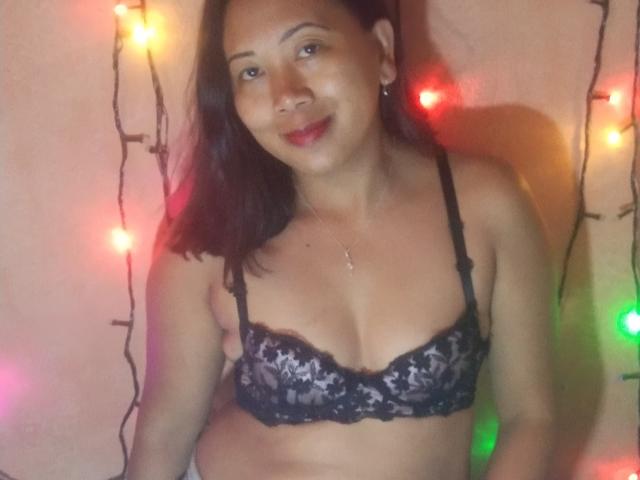 SarahFontaineX with dark brown hair, medium skin, wearing a black lace bra, smiling, red lipstick, small necklace, Christmas lights in background, soft lighting, slight belly, confident expression.