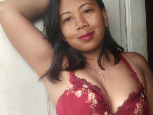 SarahFontaineX with dark hair, red lace bra, floral patterns, medium tits, arm raised, silver necklace, pink lips, slight smile, against white wall, soft natural light.