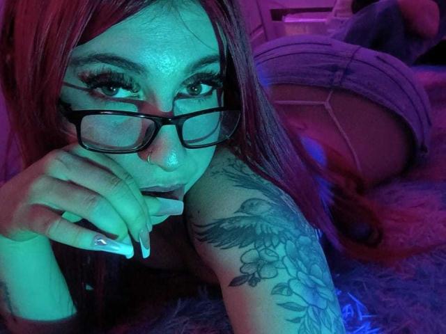 SammyViolet with red hair, glasses, and tattoo on right arm, lying on purple and blue lit furry rug, showing off her ass in shorts, looking at camera.
