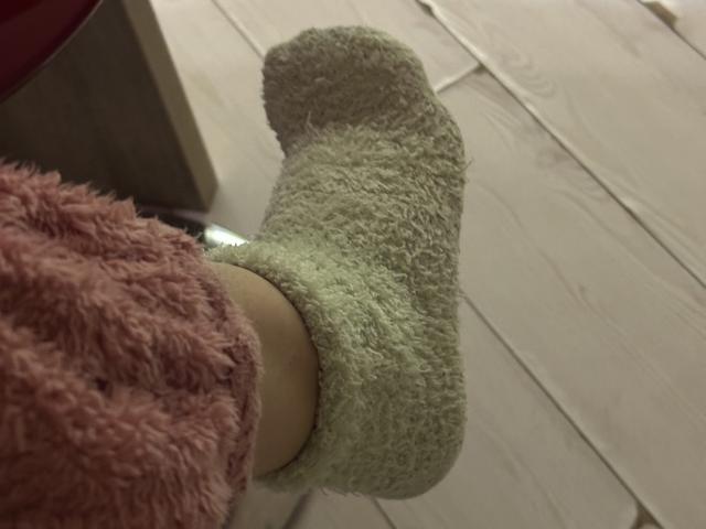 Vinnlove's foot is in a green, fuzzy sock, with a pink, fluffy sweater. Wooden floor and a dark object are in the background. Slightly blurry image.