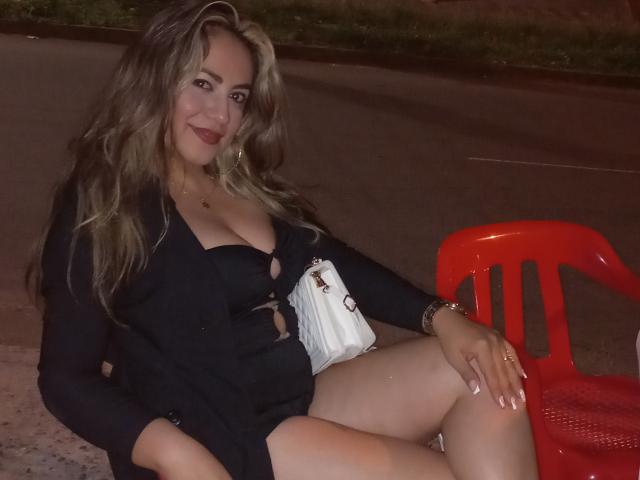 AmandaLeon with blonde hair, black dress, cleavage, red chair, white purse, sitting on pavement at night, smiling, legs crossed, wearing gold bracelet and ring.