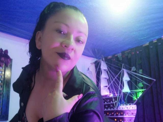 CharloteEzra with dark purple lipstick, black leather jacket, and wet hair, touching her chin, showing her small tits, in a blue-lit room with a metallic structure.