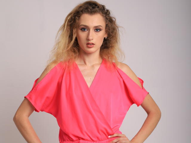 RinaPinkThree with curly blonde hair, wearing a bright pink, off-shoulder dress, standing with hands on hips, looking directly at camera, neutral expression, subtle makeup.