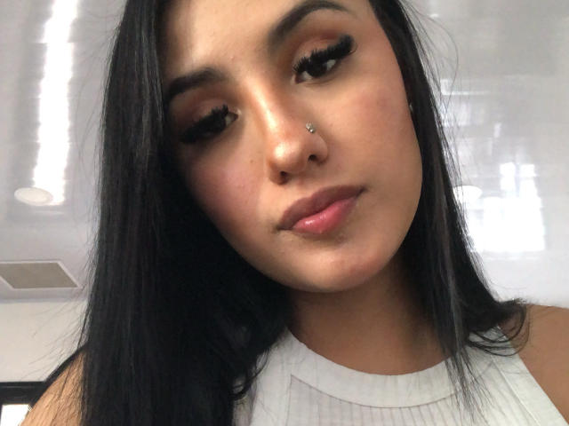 NickiClark has long black hair, a nose piercing, and full lashes with thick mascara. She's wearing a white sleeveless top, and her pink lips are slightly parted.