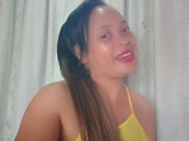 Rahel69 has shiny skin, smiling with red lips, wearing a yellow spaghetti strap top, and has long brown hair with a slight wave. Her tits are small.