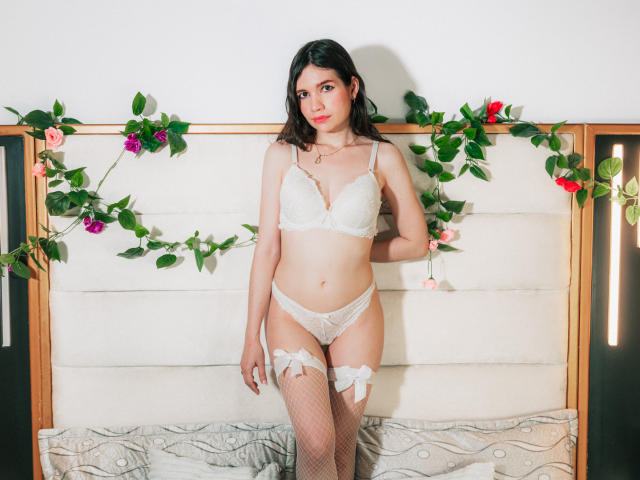 SofiArias with medium tits, white lace bra, panties, and garter, standing on bed, wearing fishnet stockings, floral wall decor, and a necklace, looking at camera, smiling.