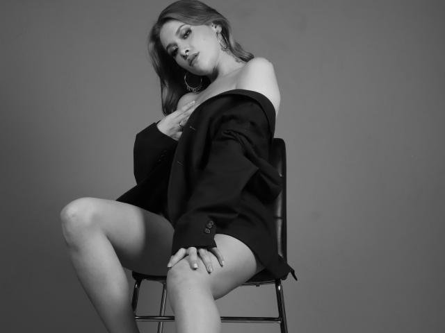 SweetAbey, assise sur une chaise, portant une veste noire à une épaule, dévoilant ses seins, les jambes écartées, regardant vers le bas, photo en noir et blanc. Sa main touche sa poitrine, une pose sexy..