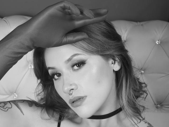 Une main gantée de noir repose sur la tête de SweetAbey, qui porte un anneau de nez, un maquillage discret et un collier ras du cou, avec des tatouages visibles sur son épaule. Elle regarde l'appareil photo..