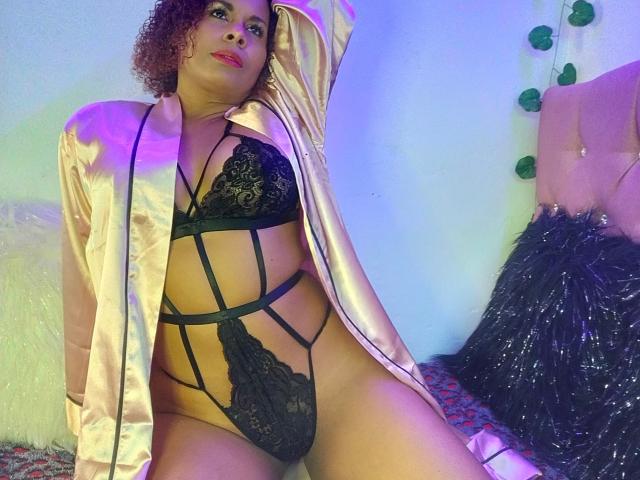 AfroditaKane, com cabelos castanhos cacheados, vestindo uma túnica rosa brilhante e lingerie de renda preta, expondo sua vulva e seios, sentada com um braço atrás da cabeça..