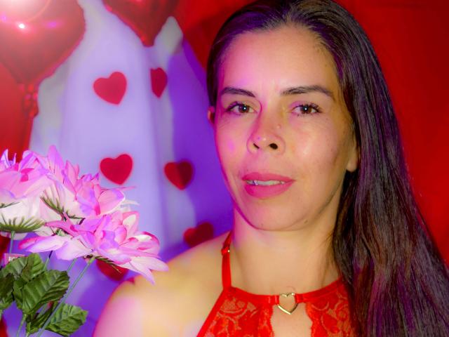 MarilynDavis with pink flowers, wearing a red lace halter, has long brown hair, light skin, and a subtle smile, her red heart necklace visible, looking cute and sexy.
