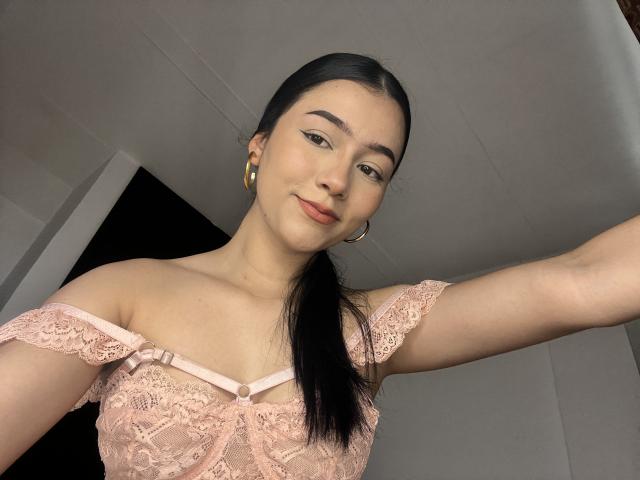 DeisyWest with black poneytail, wearing pink lace off-shoulder top, gold hoop earrings, smiling slightly, pale skin, small tits, subtle makeup, arm raised.