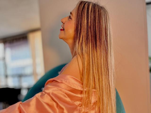 VictoriaJhonnes with long blonde hair, pink satin shirt off shoulders, smiling sideways, exposed upper chest, soft light, relaxed pose, teal chair, warm sunlight, casual, elegant.