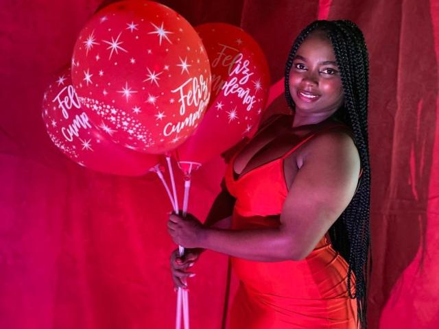 SamyLorenss, con trenzas largas, vestida con un vestido rojo, sosteniendo tres globos rojos con patrones de estrellas blancas y texto, sonriendo, mostrando su cuerpo curvilíneo, de piel oscura y voluptuoso..