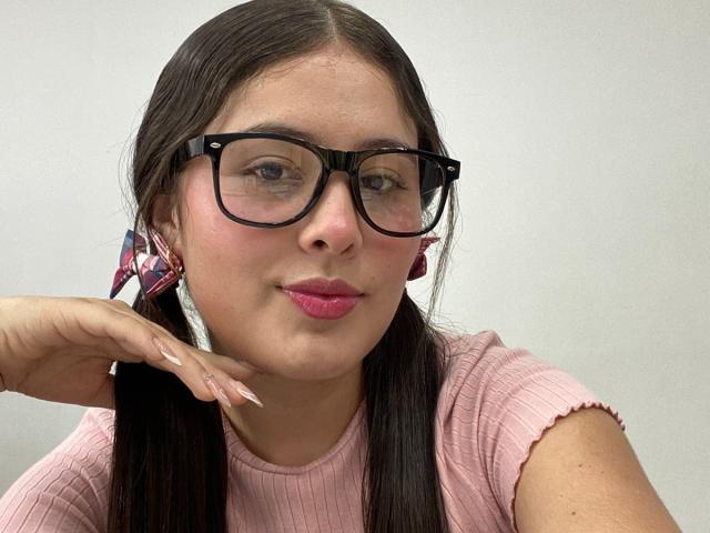 Kathy Stone, con coletas, gafas negras, lápiz labial rosa, una camisa rosa con textura y aretes rosa a cuadros, tiene una cara linda y uñas largas y cuidadas que descansan sobre su mejilla..