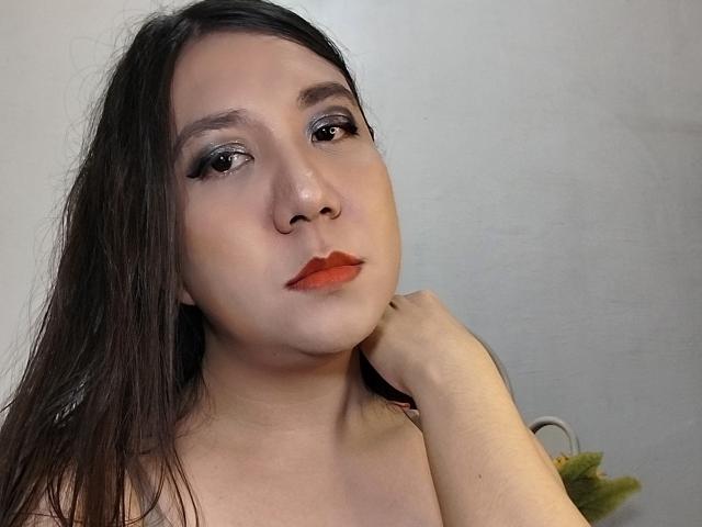 Stella, con un cabello castaño largo, usando lápiz labial rojo, delineador de ojos negro, y mirando ligeramente hacia un lado, con la mano en el cuello, piel pálida, fondo blanco, expresión suave..