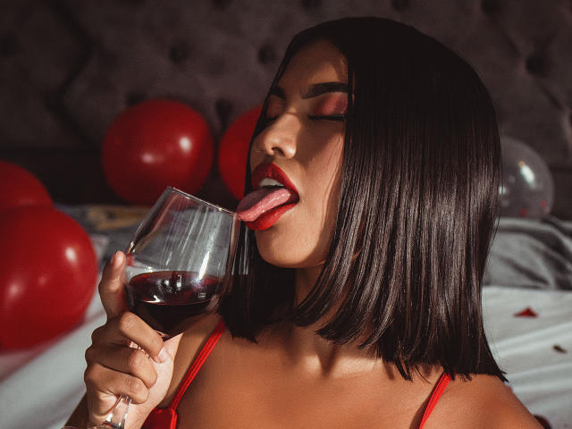 KimJohnsson avec une coupe de cheveux courte noire, rouge à lèvres rouge, la langue tirée, léchant du vin rouge dans un verre, portant de la lingerie rouge, tenant un verre de vin foncé, des ballons rouges en arrière-plan..