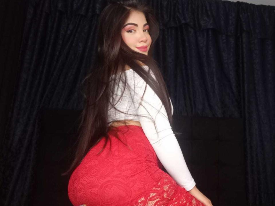 AnnaMar with long dark hair, white top, and red lace skirt, showing her big ass and curvy butt, looking over her shoulder with a slight smile.