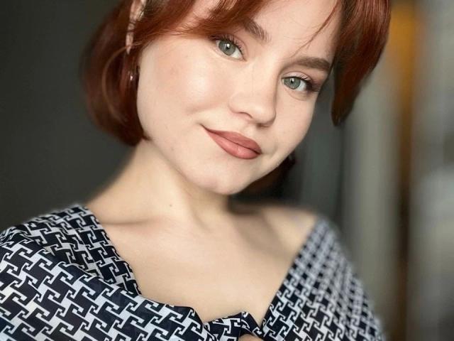Lumuon-hot heeft rood haar, groene ogen en ze glimlacht met roze lippenstift. Ze draagt een zwart-wit gestreept top, die haar borsten en billen laat zien op de foto..