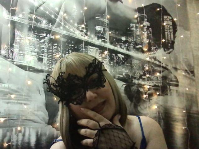 CarlySweetie with blonde bob, wearing black lace mask, fishnet glove, blue bra, hand on chin, string lights, black-and-white cityscape backdrop. Boobs, mask, glove, bra visible.