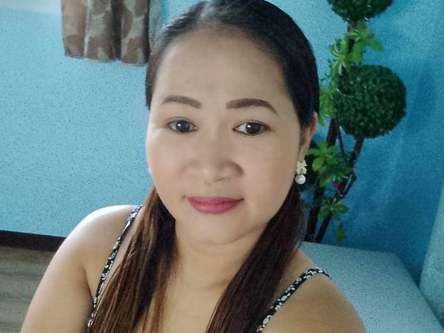 SimplyPinay with brown pigtails, wearing a black and white patterned top, has pink lipstick, small earrings, and a slight smile. Her brown eyes are slightly open.