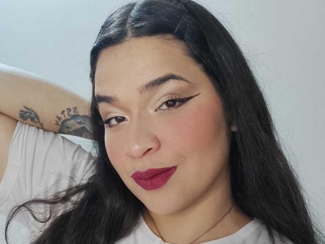 AntonelaPark has long black hair, brown eyes with winged eyeliner, red lipstick, and a tattoo on her left arm. She's wearing a white shirt, looking at the camera.