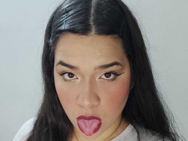AntonelaPark with long black hair, black headband, and white shirt, sticking out pink tongue, looking into camera with brown eyes, thick eyebrows, and red lipstick.