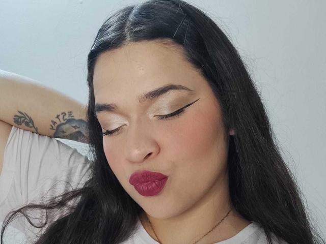 AntonelaPark with closed eyes, dark long hair, pinkish-red lipstick, white makeup, and a tattoo on her arm, pouting her full, round ass. Wearing a white shirt.