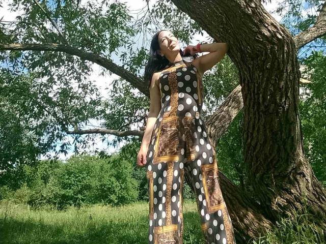 MirandaHott69 stands against a tree, wearing a brown polka dot dress, her ass slightly visible, dark hair, arm raised, green grass, sunny day, tree trunk on right.