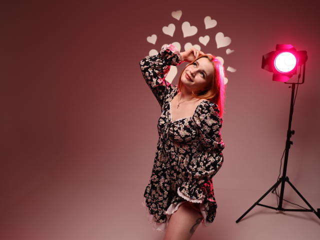 MargaritaKis with red hair, wearing a floral dress, smiling, hand in hair, standing by a red light, heart patterns on wall, showing her ass, tattoo on thigh.