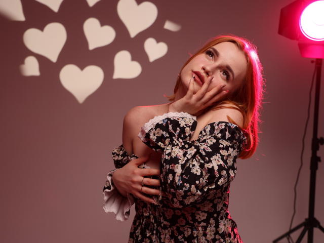MargaritaKis met rood haar, gekleed in een bloemenjurk met een open schouder, die haar gezicht aanraakt met haar hand voor haar mond, staat naast een rood licht, met harten die op de muur geprojecteerd worden..