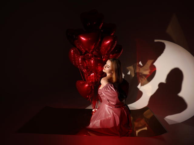 MargaritaKis in pink off-shoulder dress holding red heart balloons, sitting with back to camera, shadow of crescent moon and her ass on dark floor.