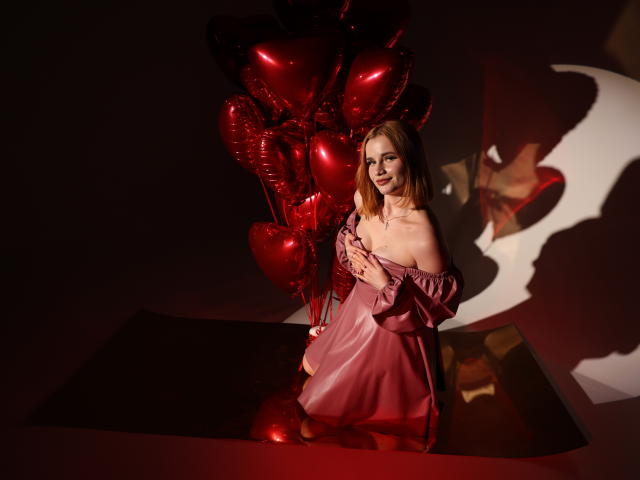 MargaritaKis with red heart balloons, wearing an off-shoulder pink dress, standing in front of a shadowy wall, looking at the camera with a subtle smile.