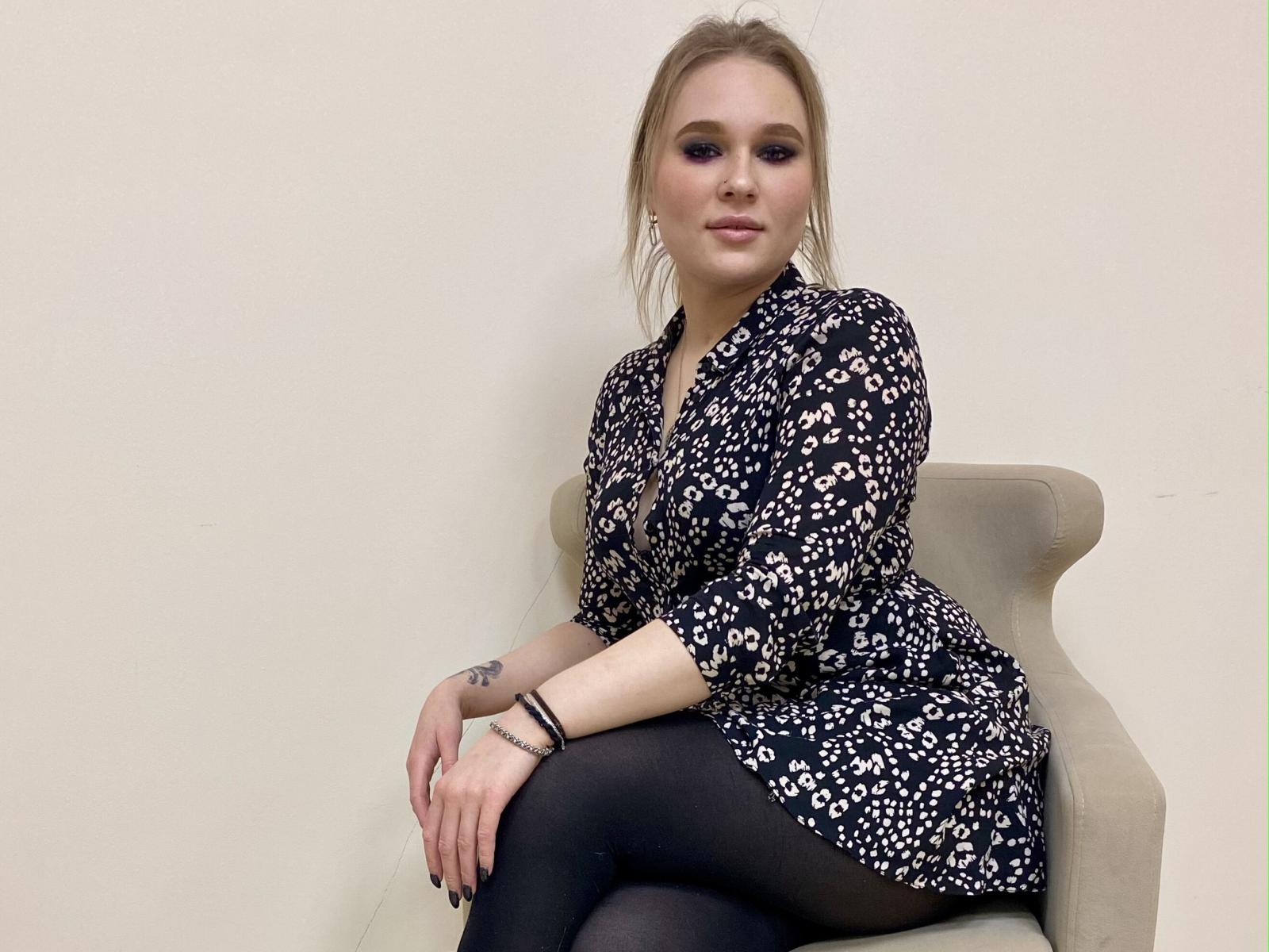 CloeHarris with blonde hair, wearing a black floral dress, black tights, sitting on a beige chair, hands on knees, tattoo on left wrist, subtle makeup, confident smile.