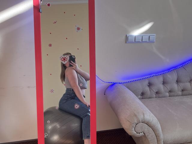 DiLaria, avec ses fesses moulées dans un jean ajusté, portant un masque fleuri, est assise sur une boule grise devant un miroir à bordure rouge, à côté d'un canapé beige capitonné..
